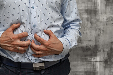 Close-up of a man's hands holding his aching stomach. A man wearing a blue long-sleeved shirt stands in a medical clinic