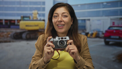 Woman wearing brown jacket and yellow shirt smiling while holding vintage camera at a building site under natural light; curiosity exploration.