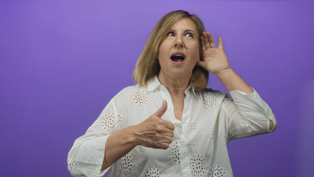 Middle age woman cups hand to ear with thumb up in a purple studio setting to show listening engagement; attentiveness.