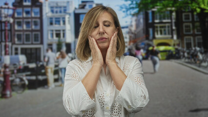 Fototapeta premium Woman presses her cheeks with both hands on a sunny european street lined with bicycles and canal houses; thinking.