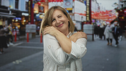 Fototapeta premium Woman hugs her shoulders on bustling street scene with blurred passersby and city lights; self love contentment.
