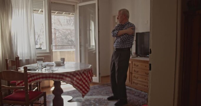An elderly man in his home attempts to stretch and alleviate intense back pain. The discomfort highlights daily challenges faced by older adults maintaining physical health and mobility.