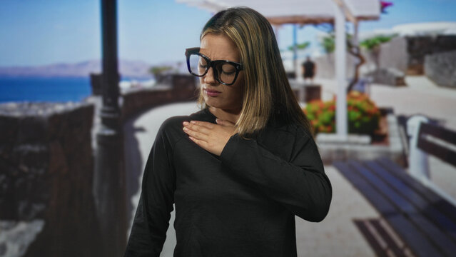 Woman touching shoulder on a seaside street near bench and stone railing, wearing black top and large glasses with closed eyes showing pain; discomfort travel fatigue.