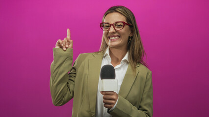 Woman reporter holding microphone points finger and laughs broadly in pink studio while wearing olive blazer; enthusiasm.