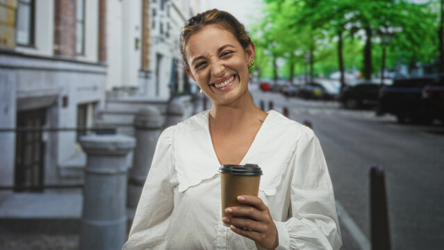 Woman smiling and holding a takeaway coffee cup while walking down an urban street lined with parked cars and trees; morning joy.