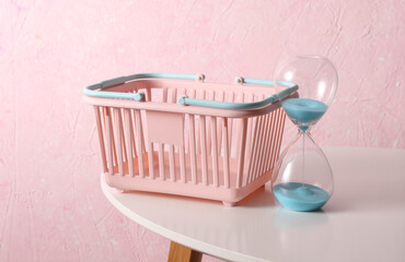 Supermarket basket and Hourglass on table, pink concrete background. Shopping time