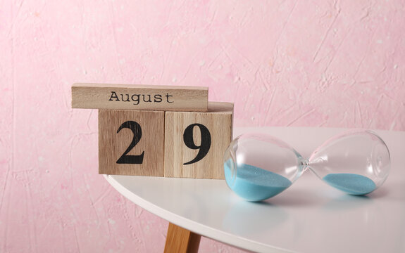 August 29 wooden block calendar and hourglass on table, pink concrete background - Powered by Adobe
