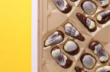 Box of chocolate candies in the shape of sea shells on yellow background. Top view