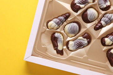 Box of chocolate candies in the shape of sea shells on yellow background. Top view