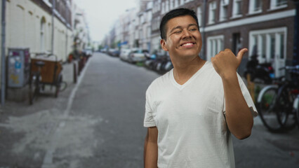 Fototapeta premium Hispanic young man pointing thumb over shoulder on street while smiling; casual confidence happiness.