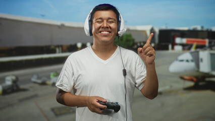Young hispanic man with headset holds game controller and points finger upward in airport terminal; joyful play.