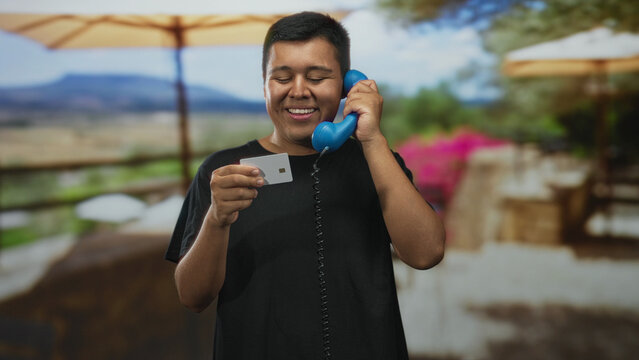 Man holding creditcard and blue handset to ear while smiling on street terrace at outdoor restaurant under umbrella; relaxed contentment.