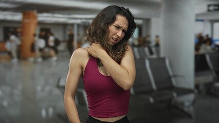 Woman holds shoulder in busy airport terminal with tense expression and slightly furrowed brow; discomfort.
