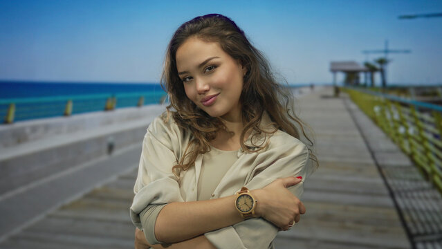 Woman hugging herself wearing a watch while standing on a wooden seaside promenade under bright daylight; self care calm serenity.