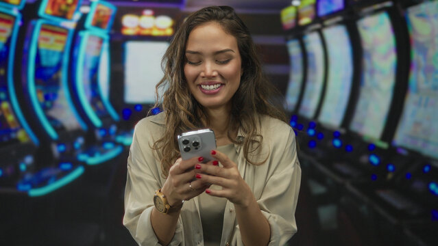 Young hispanic woman at casino slot machine laughs while texting on smartphone indoors, conveying enjoyment and connectivity in a lively gaming environment.