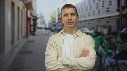 Man standing confidently on an urban street with arms crossed, wearing casual beige shirt amidst city environment, showcasing relaxed outdoor vibe in daylight.