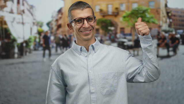 Man smiling with glasses and striped shirt giving thumbs up on bustling street with people and historical architecture in background on sunny day, showcasing urban life charm. - Powered by Adobe