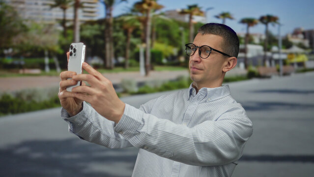 Man using smartphone on outdoor street with palm trees and blurred background, capturing a casual moment in public space, highlighting communication and technology.