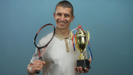 Man smiling holding tennis racket and trophy against isolated blue background showcasing achievement in sports.