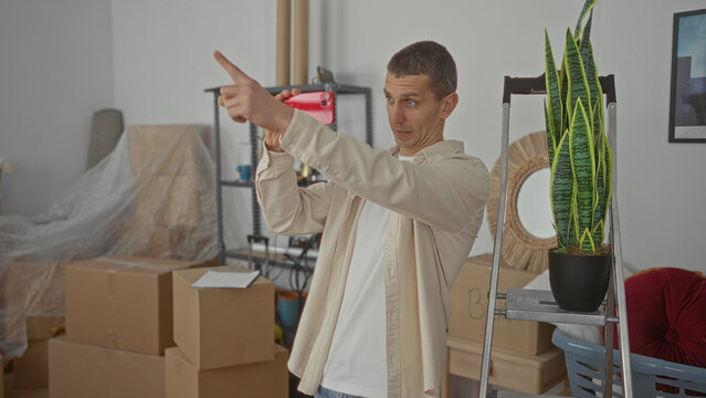 Man recording video with smartphone in living room surrounded by moving boxes and houseplants, creating a personal vlog in his new home reflecting a fresh start and creativity.