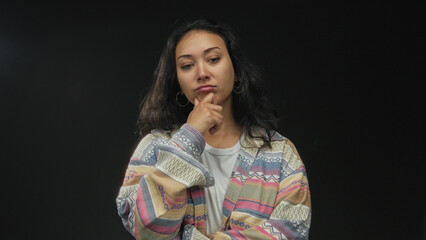 Fototapeta premium Woman with hand on chin in black studio wearing patterned cardigan and hoop earrings; thoughtful introspection.