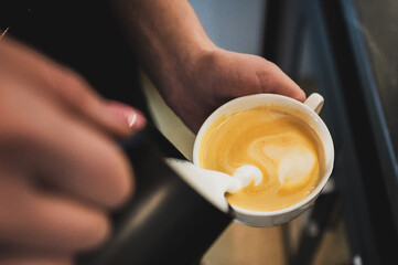 Close-up of a barista's hands pouring steamed milk into a white ceramic cup to create latte art, focusing on the creamy texture and action of coffee preparation