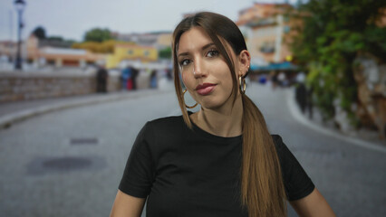 Woman tilts head to feature her face and gazes at camera on a sunlit city street wearing a black...