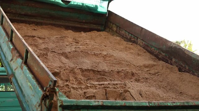 Close up of a rusty turquoise dump truck bed tilted upward as coarse tan sand shifts, showing ridged texture, corroded panels, and green tarpaulin in daylight