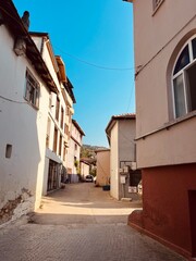 A sunlit photograph of a narrow, cobblestone street lined with pastel-colored buildings in a quaint European town. The sky is bright blue with a few power lines visible.