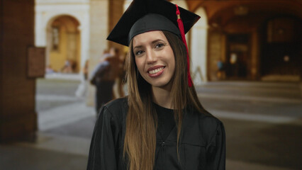 Young hispanic woman wearing graduation cap and gown smiles with teeth visible in front of building; achievement pride.