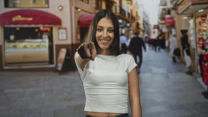 Young hispanic woman points finger at camera on street smiling and engaging with viewer; confidence connection.
