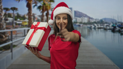 Obraz premium Woman holding gift and extending hand on street pier wearing santa hat and red shirt; holiday gift giving joy.