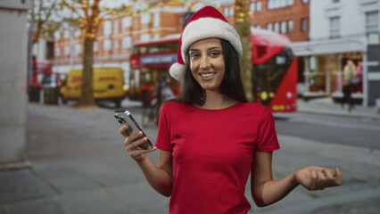 Woman holding smartphone and smiling on city street in red santa hat and red shirt; holiday cheer warmth.