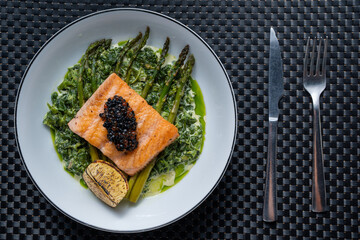 Grilled salmon fillet topped with black caviar served on a bed of asparagus and creamy spinach with a lemon wedge on a white plate