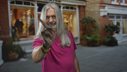 Man with long hoary hair and beard pointing index finger to camera on street near lit shop windows and potted plants at night; defiance.