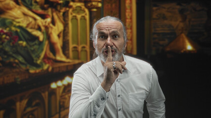Man with long gray hair and beard points finger to lips for silence in gallery building; quiet contemplation.