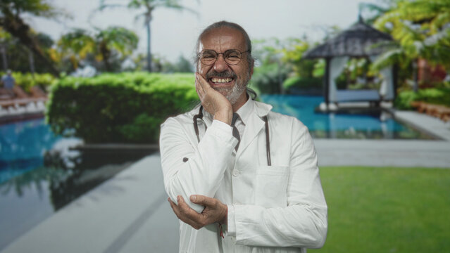 Man doctor smiling with hand on cheek and stethoscope poolside by resort building; compassionate care joy.