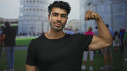 Young hispanic man flexes arm muscle with clenched fist and slight smile at pisa tower building; confidence.