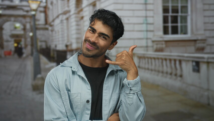 Obraz premium Young hispanic man makes playful call me gesture with hand in a busy city street setting; friendly invitation.