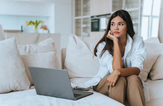 Woman comfortably seated with an expression of contemplation, leaning on one hand, beside an open laptop, capturing a moment of introspection.