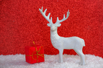 A charming Christmas reindeer figure stands amidst a soft layer of snow on a vibrant, shiny red...
