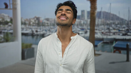 Man tilting head back with closed eyes and sunlit neck in a busy seaside port with moored boats and...