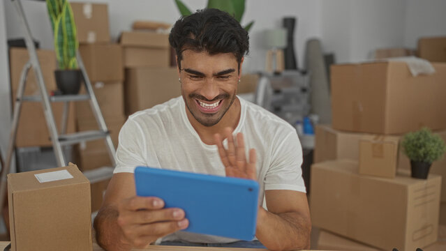 Smiling man waves hand at tablet in building surrounded by cardboard boxes and houseplant on shelf; connection.
