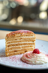 Layered honey cake slice topped with a raspberry served on a plate with cream. Elegant presentation against a blurred background.