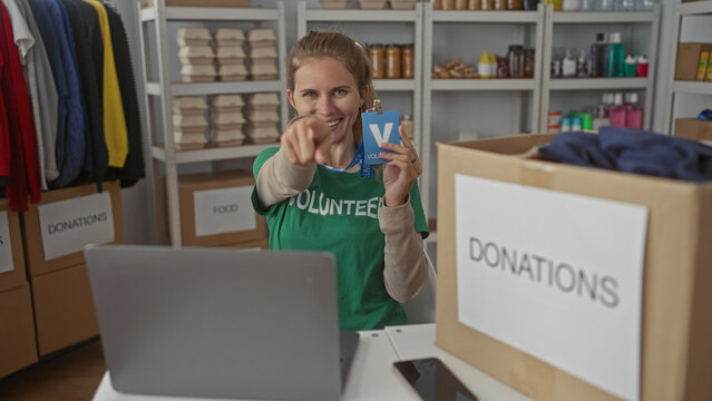 Woman displays badge and pointing gesture in center as volunteer beside laptop and smartphone, smiling with blonde young features.