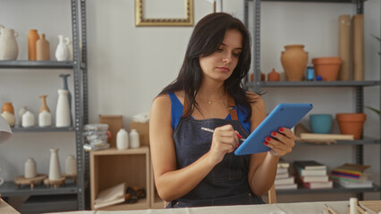 Artisan woman draws on tablet with stylus in studio amid shelves of pottery; creativity concentration.