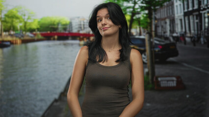 Young hispanic woman shrugs shoulders beside a canal with glisttening water on a cobblestone street; indifference.