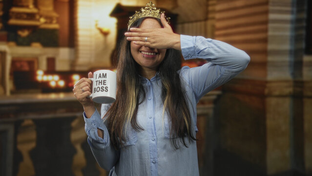 Young latina brunette woman holding a mug labeled i am the boss while covering her eyes with one hand in a church building; playful confidence.