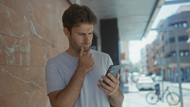 Man holding smartphone with hand on chin on street near parked bicycle and cars, looking at screen; thinking uncertainty doubt.