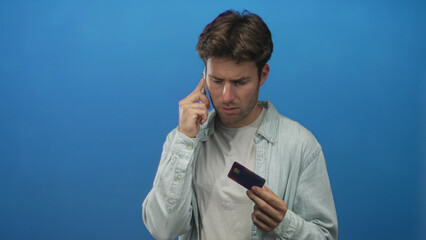 Man young hispanic frowning while holding a creditcard and smartphone to his ear and inspecting the card in a blue studio; financial concern.